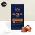 A box of CaféPod coffee pods with the Brunch Blend variety shown. The box is navy with a fox logo and copper text, and there are copper-colored coffee pods in front of the box.