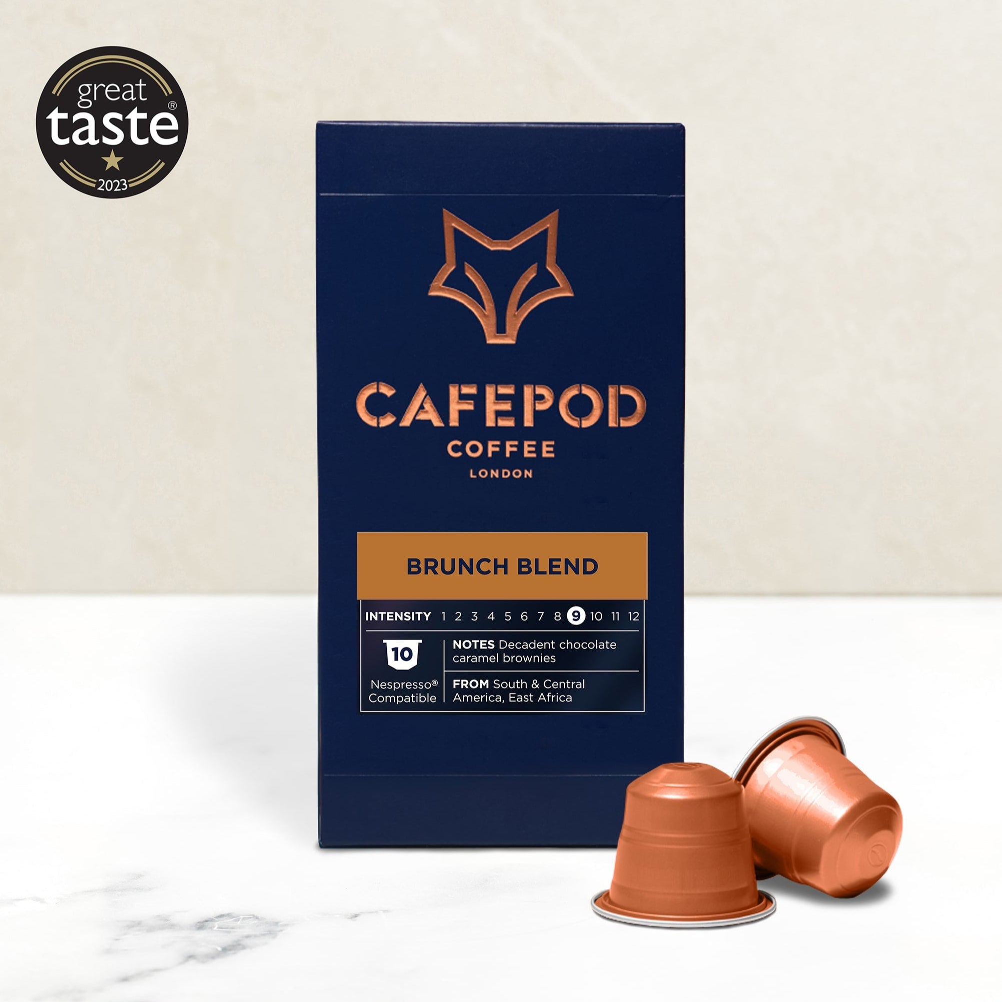 A box of CaféPod coffee pods with the Brunch Blend variety shown. The box is navy with a fox logo and copper text, and there are copper-colored coffee pods in front of the box.