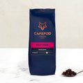A navy blue bag of CaféPod Coffee labeled 'Daily Grind Whole Bean' with a scoop of coffee beans beside it.