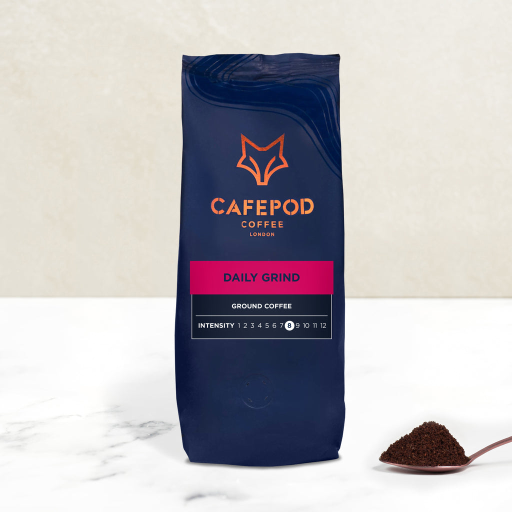 A navy blue bag of Cafepod Daily Grind ground coffee with a scoop of coffee grounds in front, against a white background.