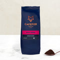 A navy blue bag of Cafepod Daily Grind ground coffee with a scoop of coffee grounds in front, against a white background.