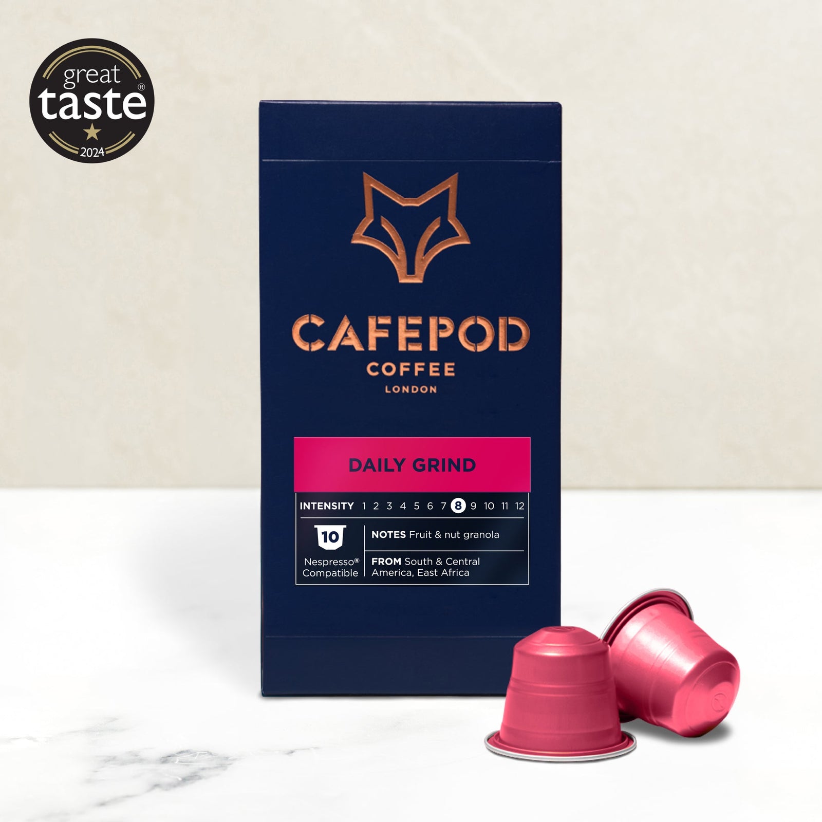A box of Cafepod Daily Grind coffee capsules with a modern design, featuring a black background and the brand's logo in gold. Two capsules are placed in front of the box, one upright and the other lying on its side.